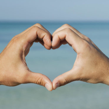 Two men's hands forming heart shape by ocean, cropped