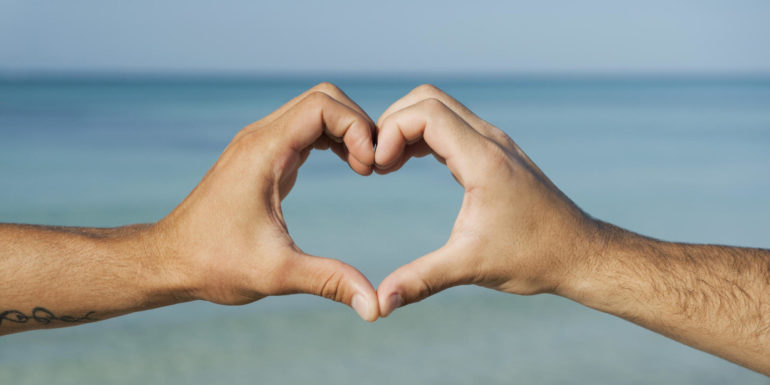 Two men's hands forming heart shape by ocean, cropped